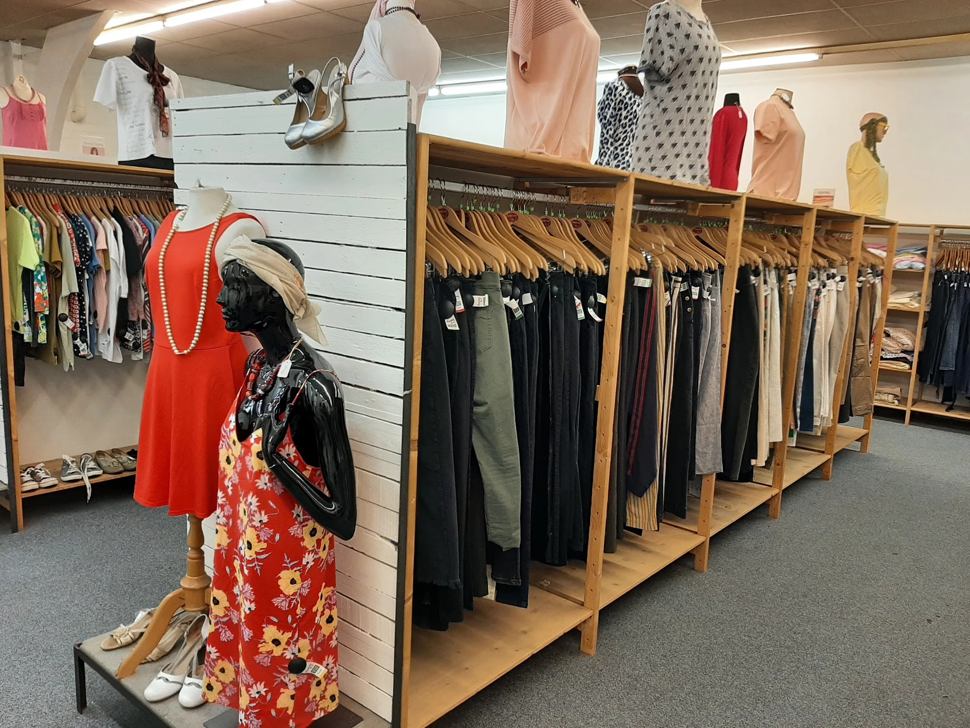 Houten rekken met uitgestalde dameskleding, waaronder jurken, broeken en tops in verschillende kleuren. Een mannequin toont e