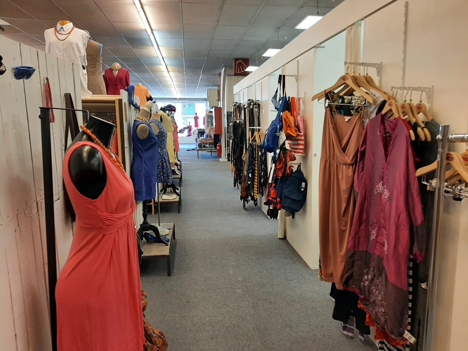 Winkelgang met kledingrekken vol tweedehands kleding in verschillende kleuren, zoals rood, blauw en bruin.