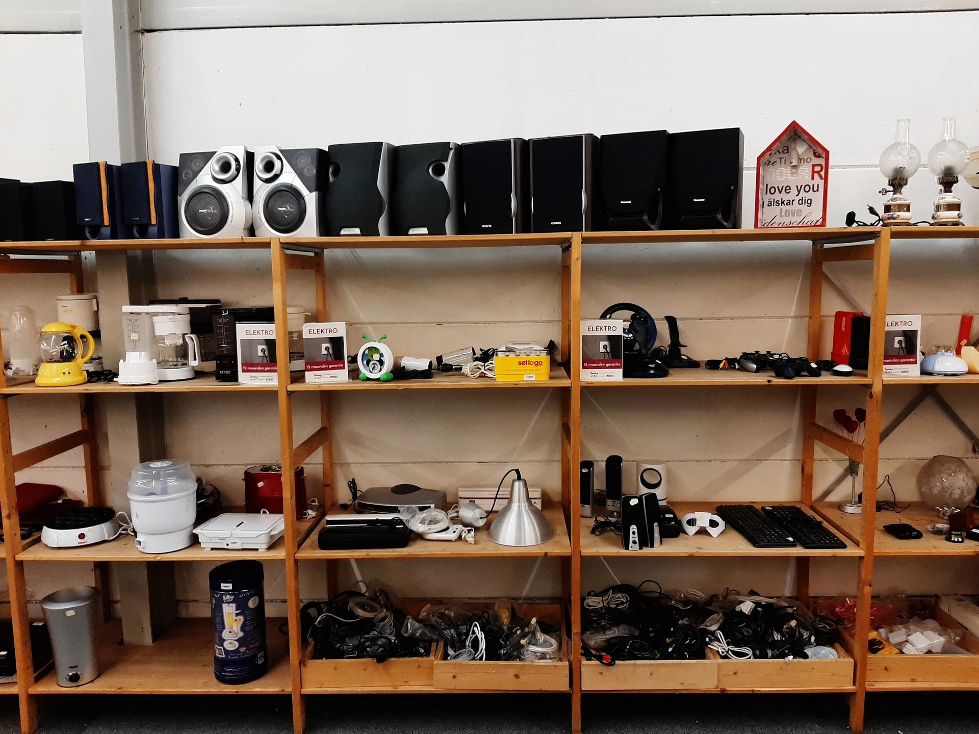 Houten stellingen met diverse gebruikte elektronische apparaten, speakers, camera's en huishoudelijke items