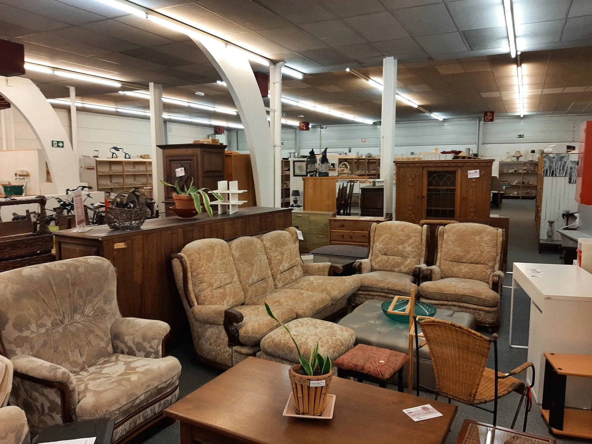 Zithoek met tweedehands meubels: fauteuils en bank in beige stof, houten kastjes en kasten op de achtergrond in een ruime win