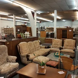 Zithoek met tweedehands meubels: fauteuils en bank in beige stof, houten kastjes en kasten op de achtergrond in een ruime win