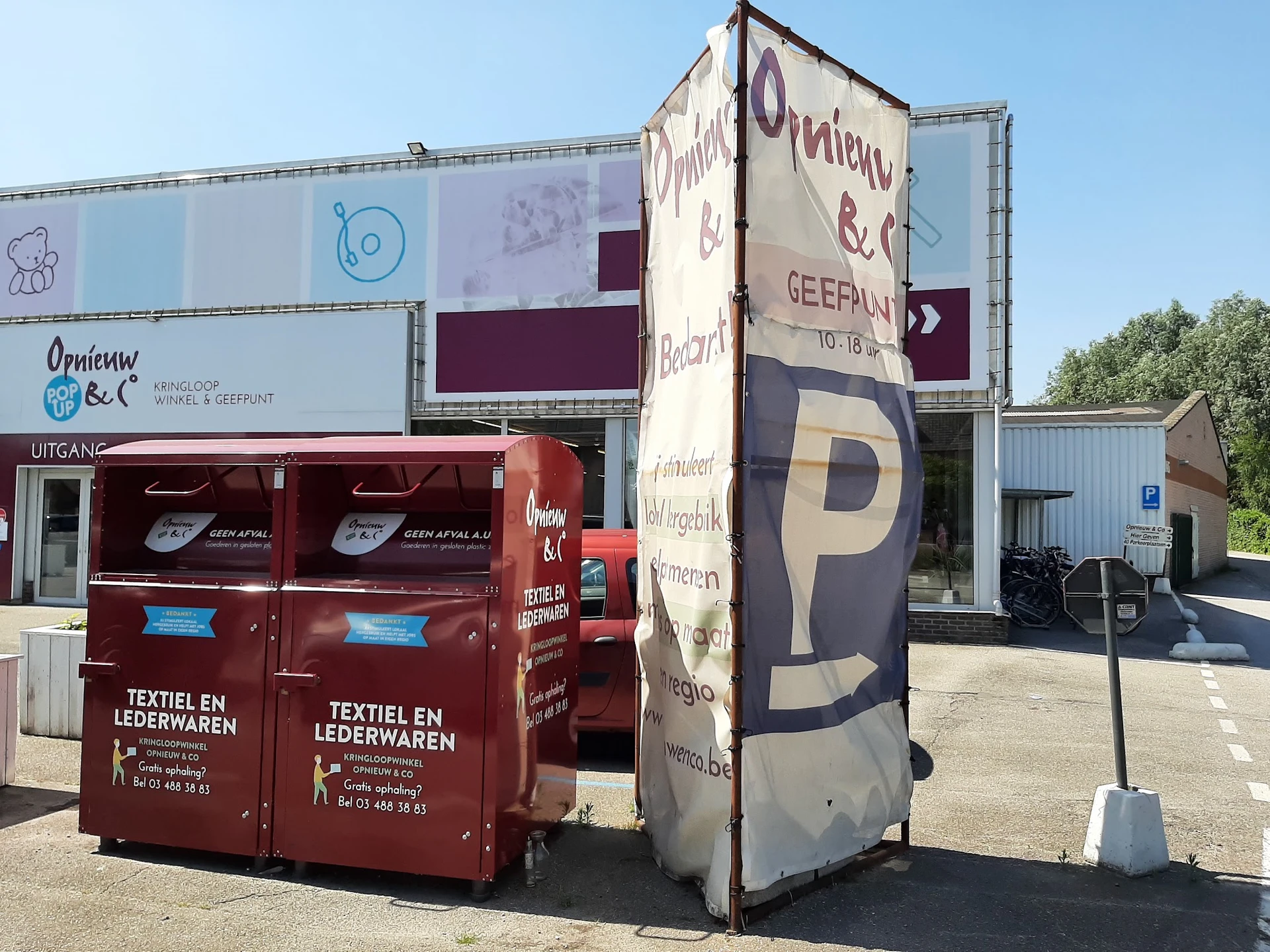 Exterieur van kringloopwinkel met rode inzamelbak voor textiel en lederwaren, parkeerplaats zichtbaar
