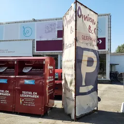 Exterieur van kringloopwinkel met rode inzamelbak voor textiel en lederwaren, parkeerplaats zichtbaar