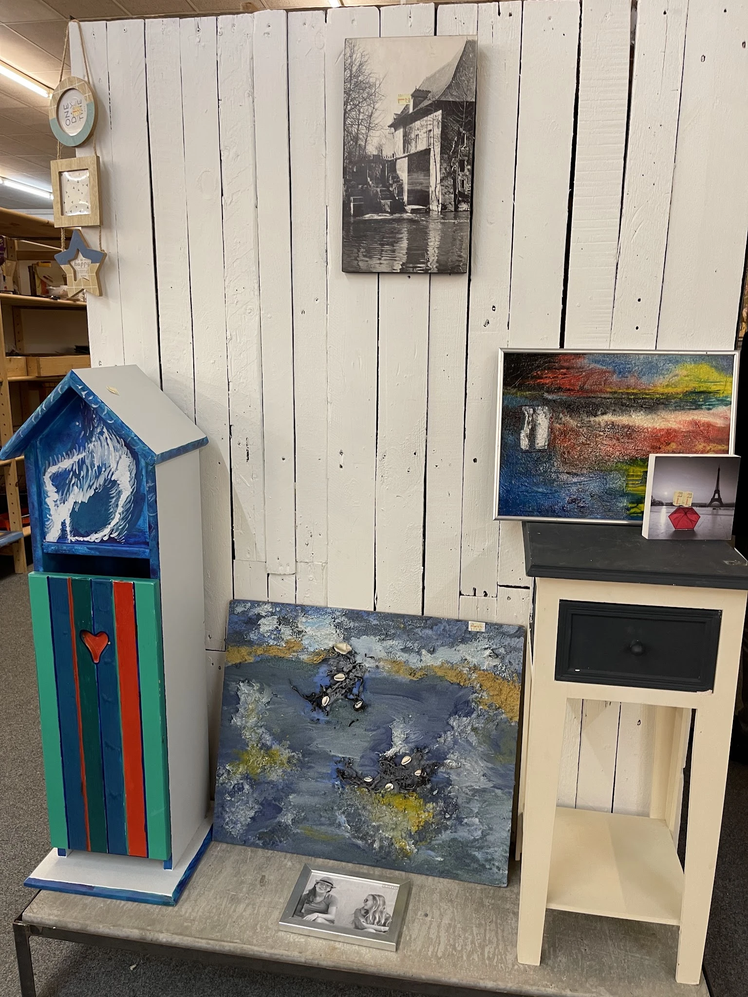 Kunstwerken en decoratieve items in de kringloopwinkel, waaronder schilderijen, een blauwe vogelhuisje en keramische kunstobj