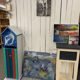 Kunstwerken en decoratieve items in de kringloopwinkel, waaronder schilderijen, een blauwe vogelhuisje en keramische kunstobj