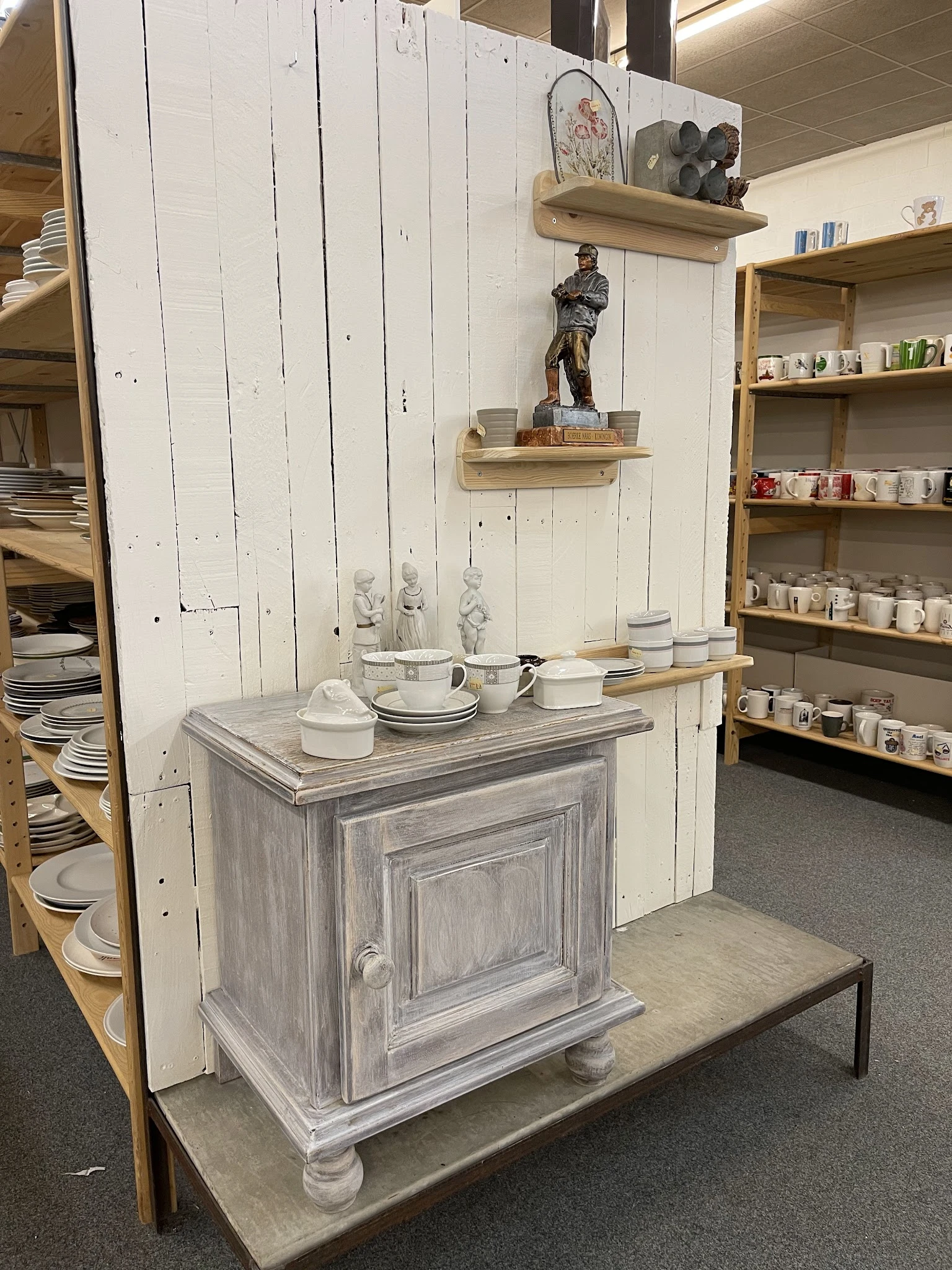 Uitgestalde serviesgoed en huishoudartikelen in een kringloopwinkel, met houten kastje, borden, kopjes en decorstukken