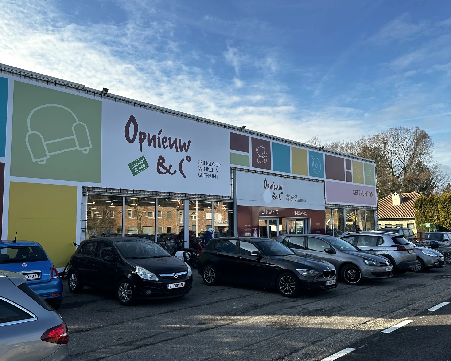 Witte gevel van kringloopwinkel met groen en geel accent, grote ramen, parkeerplaats met auto's voor de ingang