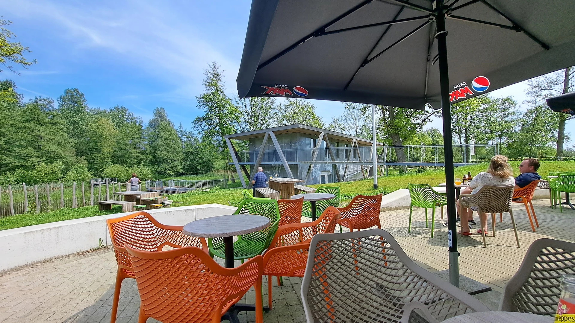 Terras met oranje stoelen onder parasol, uitzicht op speeltuin en gebouwen