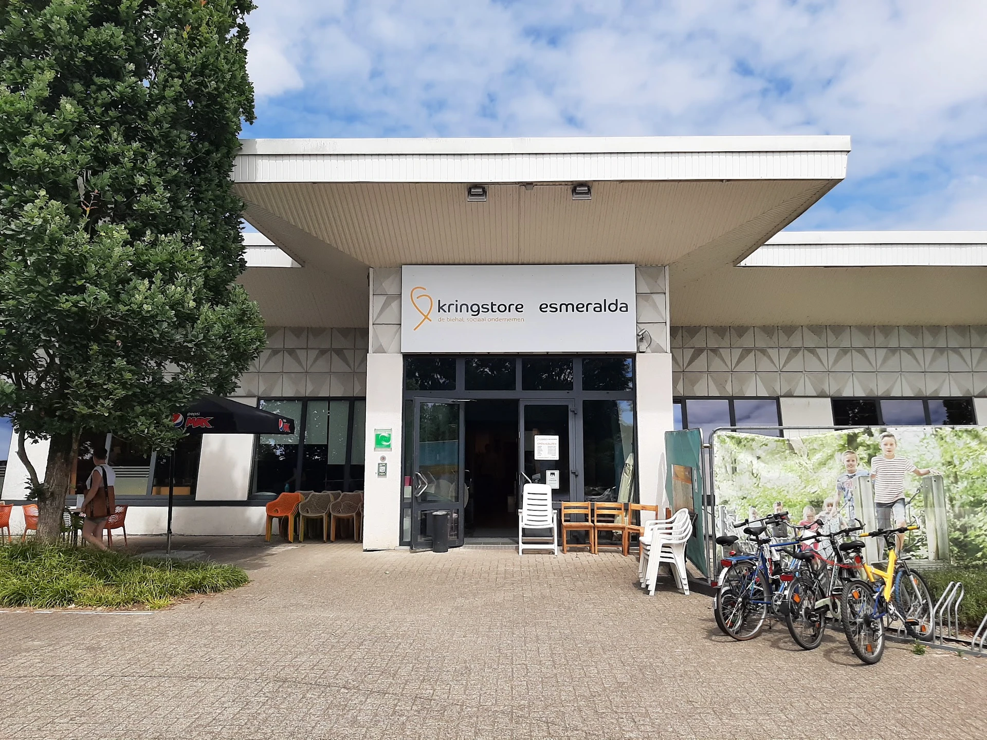Voorgevel van kringloopwinkel met witte pui, winkelnaam zichtbaar, ingang met glazen deuren, fietsen en spullen buiten