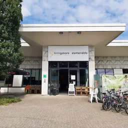 Voorgevel van kringloopwinkel met witte pui, winkelnaam zichtbaar, ingang met glazen deuren, fietsen en spullen buiten
