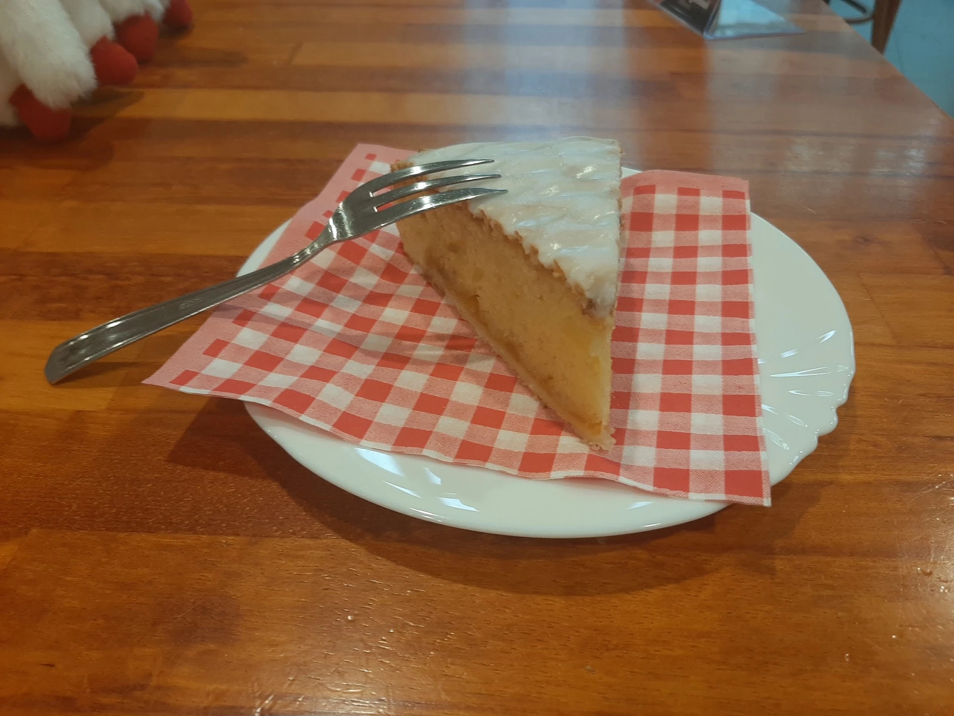 Een stuk cake met witte glazuur op een bord met rood-wit geblokt papier en vork