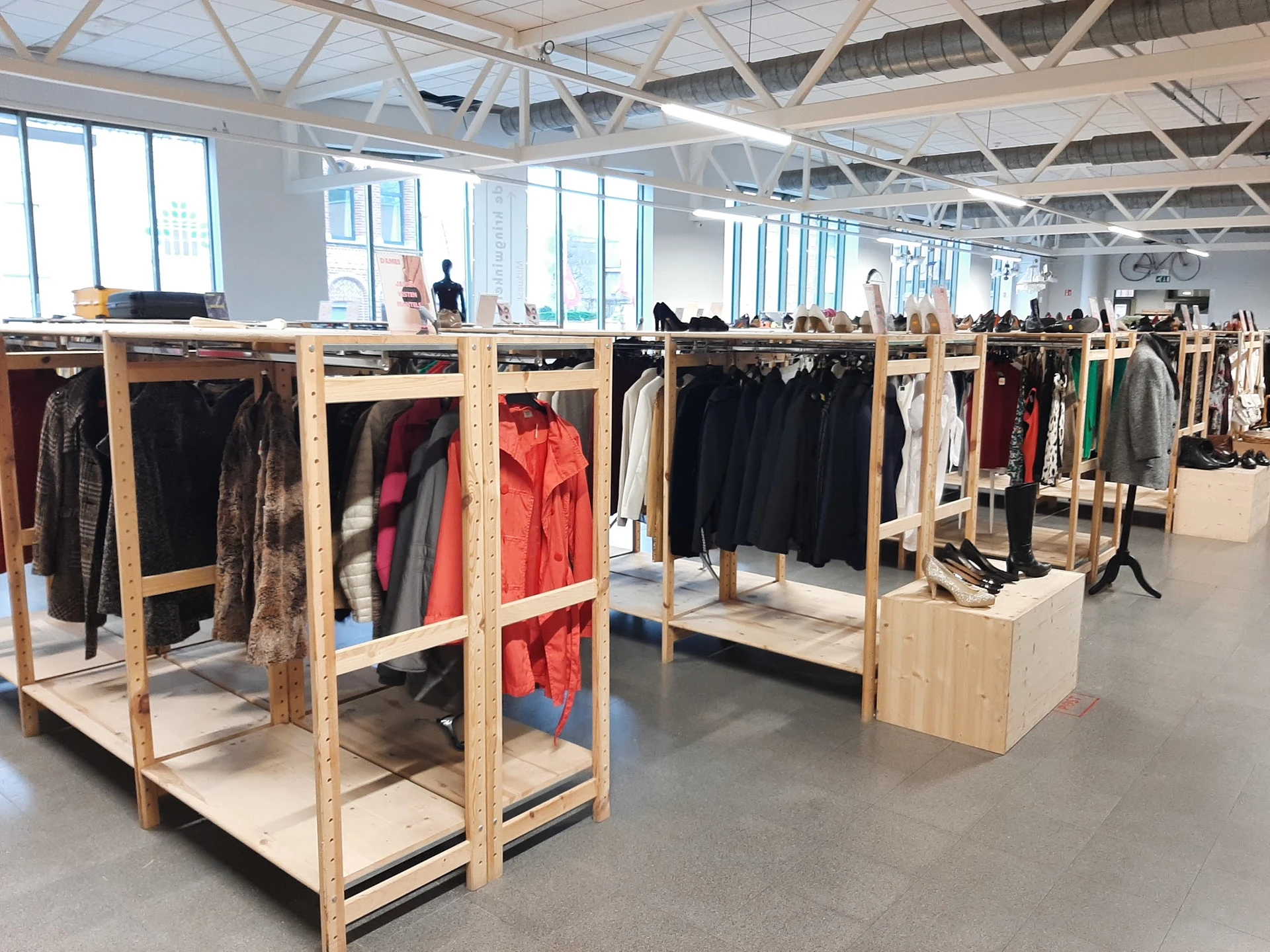Houten kledingrekken met tweedehands jassen en kleding in een ruim magazijnachtige winkelruimte.