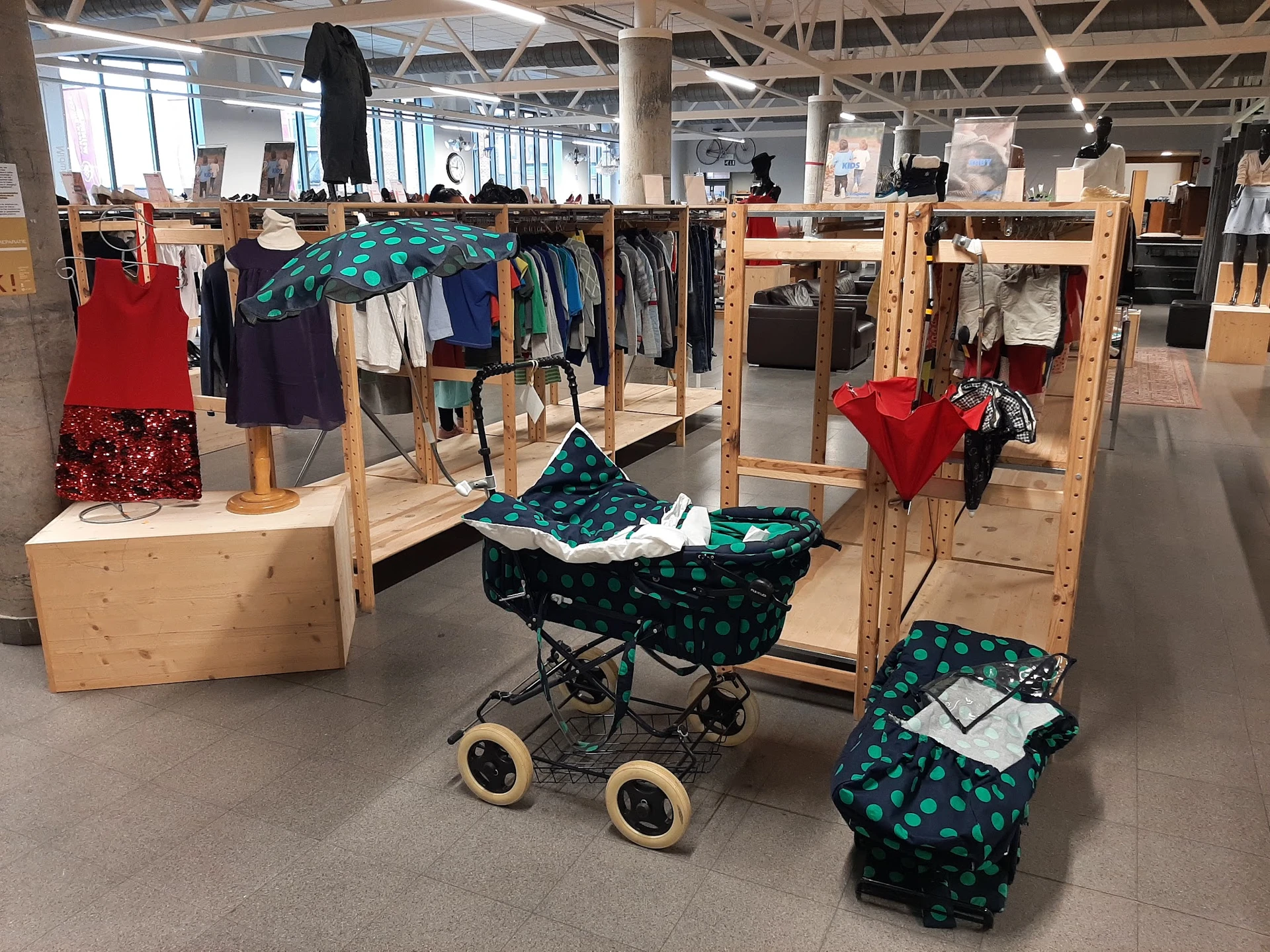 Uitgestalde tweedehands kleding op houten rekken en een kinderwagen met groene stof in een kringloopwinkel