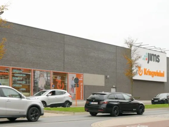 Grijze gevel van kringloopwinkel met oranje ramen en parkeerplaats met auto's ervoor