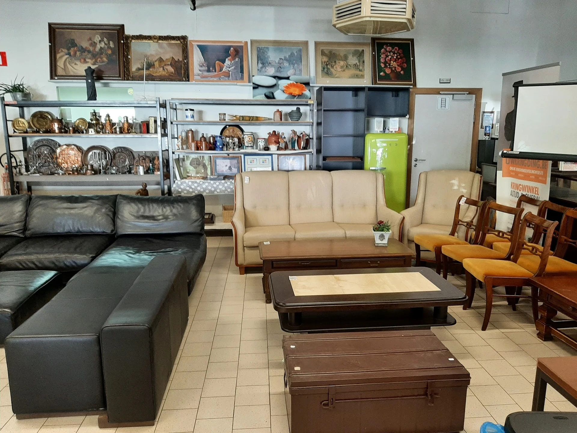 Winkelvloer met diverse meubels: zwarte leren bank, beige bank, houten stoelen, tafels en kasten met huishoudartikelen en dec