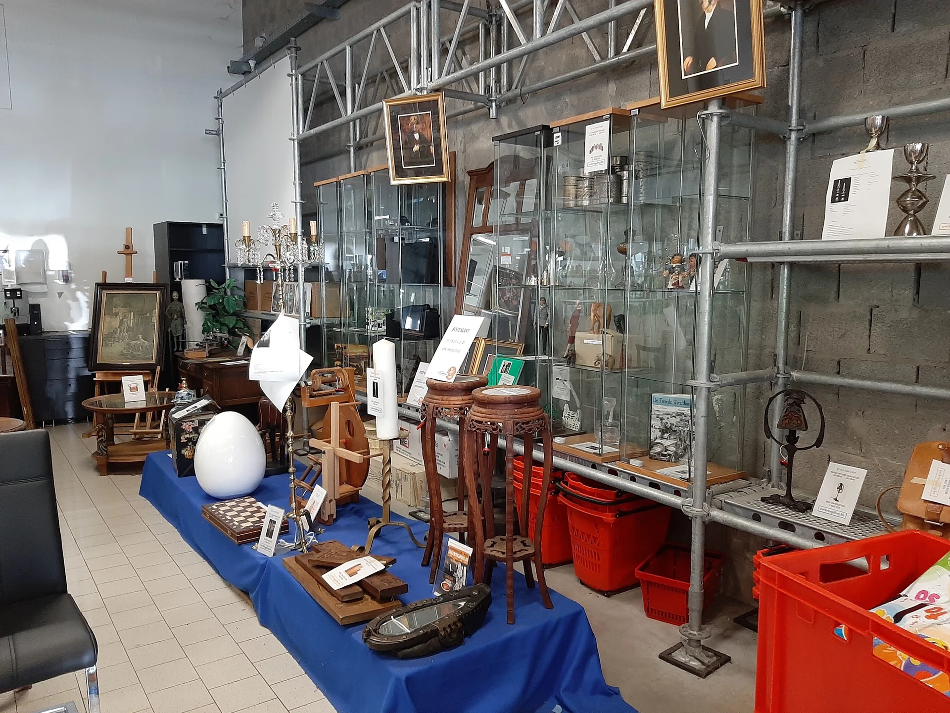 Winkelvloer met diverse tweedehandsartikelen, blauw tafelkleed en rode opbergkisten, stellingen met glaswerk en decoratieve i