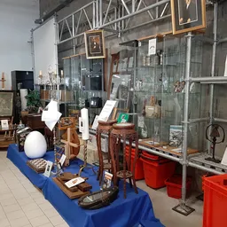 Winkelvloer met diverse tweedehandsartikelen, blauw tafelkleed en rode opbergkisten, stellingen met glaswerk en decoratieve i