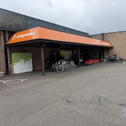 Voorkant van kringloopwinkel met oranje luifel, fietsen geparkeerd onder het afdak, bakstenen gevel