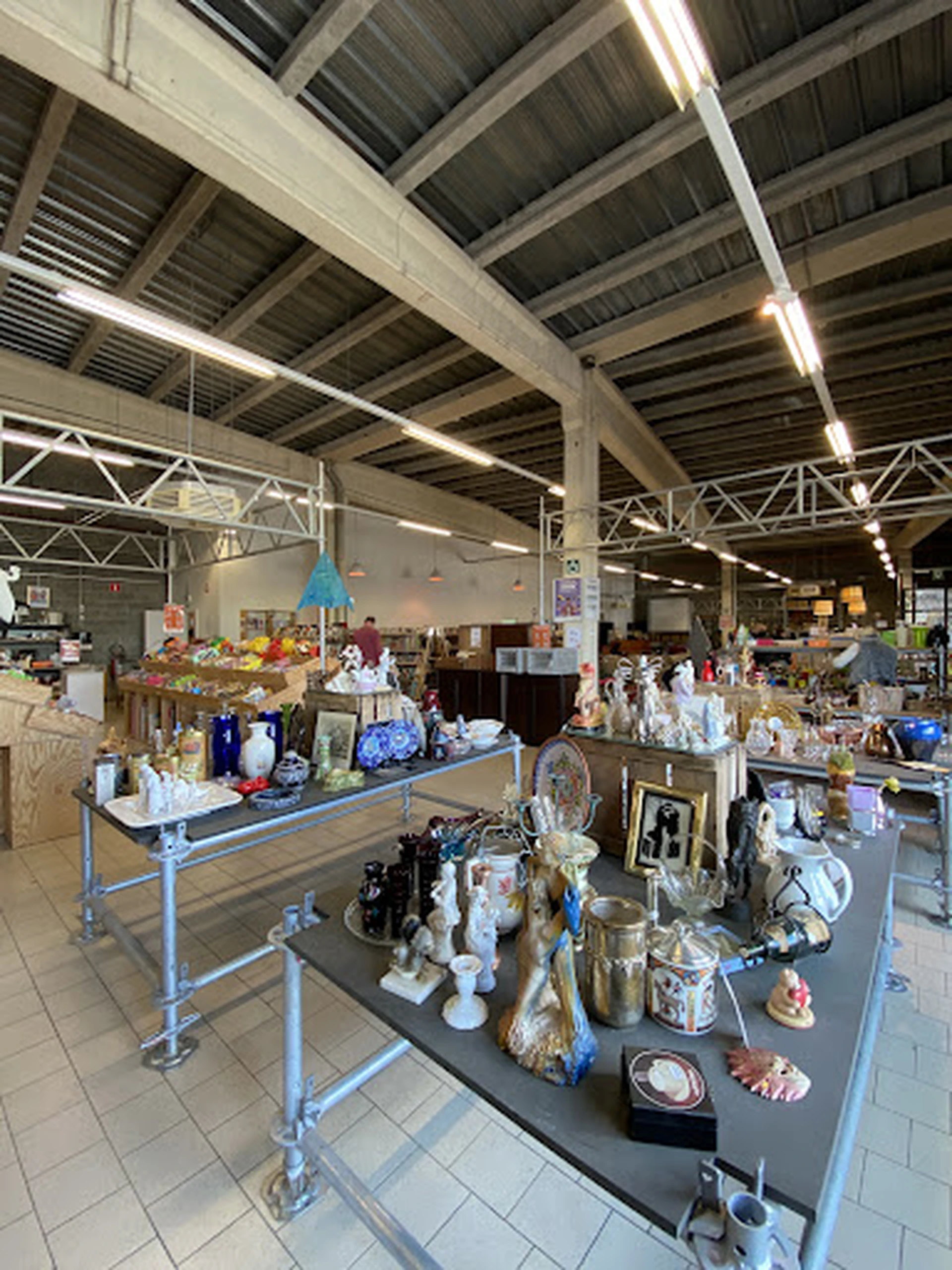 Overzicht van kringloopwinkelruimte met tafels vol decoratiespullen, servies en huishoudartikelen onder industrieel plafond