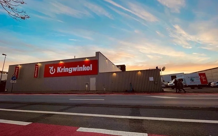 Gevel van kringloopwinkel met rode Kringwinkel-logo aan grijze metalen gevel, aan de straat met parkeerplaats