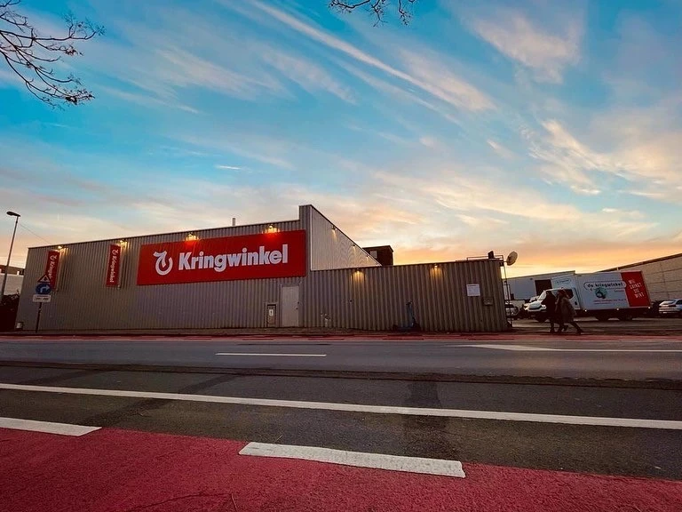 Gevel van kringloopwinkel met rode Kringwinkel-logo aan grijze metalen gevel, aan de straat met parkeerplaats