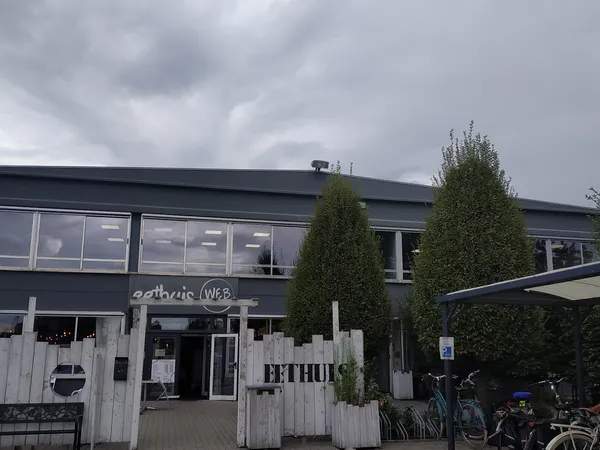Voorkant van kringwinkel met donkergrijze gevel, witte houten ingang, groene coniferen en fietsen op het terras