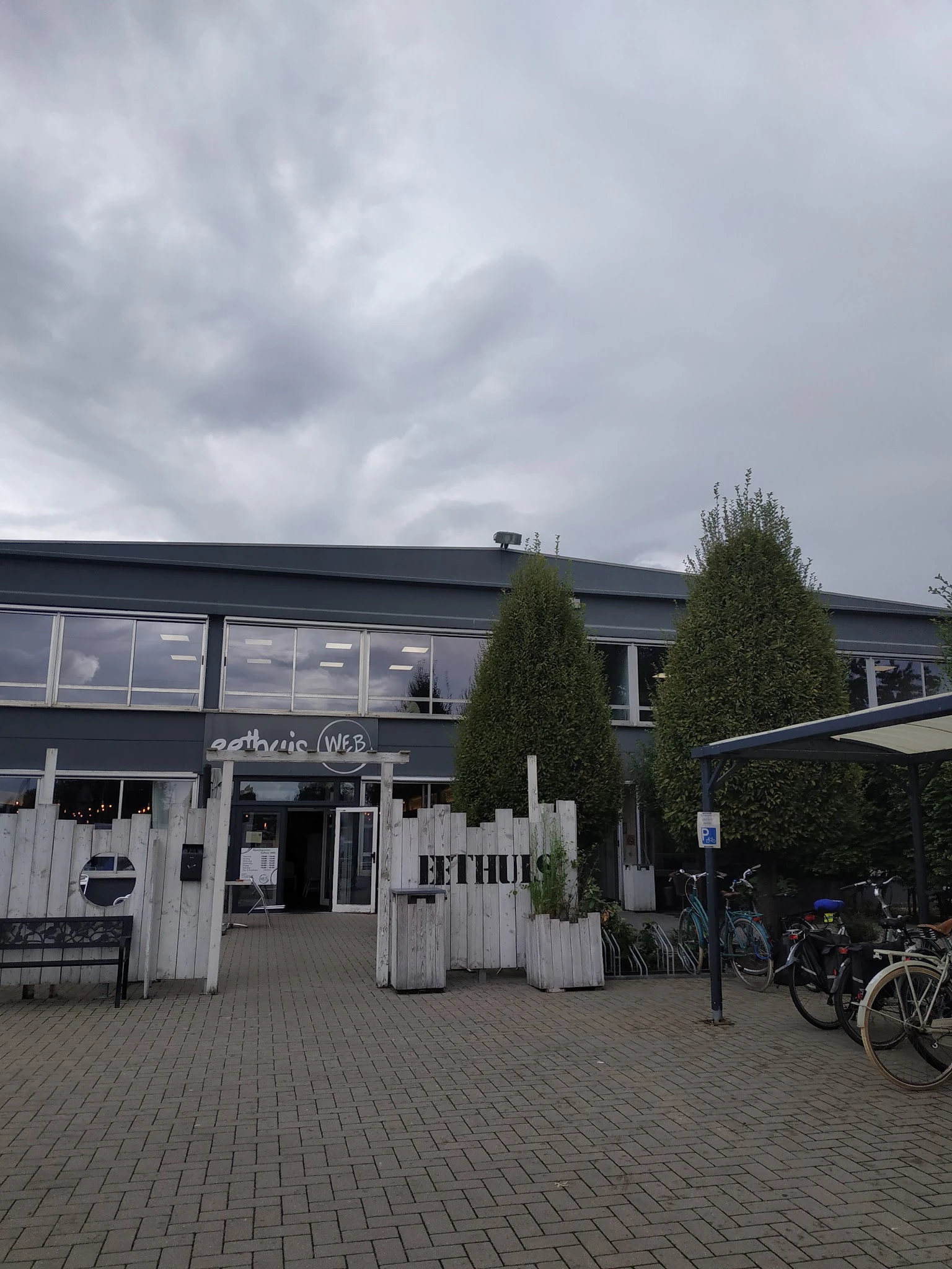 Voorkant van kringwinkel met donkergrijze gevel, witte houten ingang, groene coniferen en fietsen op het terras