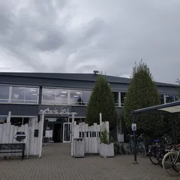 Voorkant van kringwinkel met donkergrijze gevel, witte houten ingang, groene coniferen en fietsen op het terras