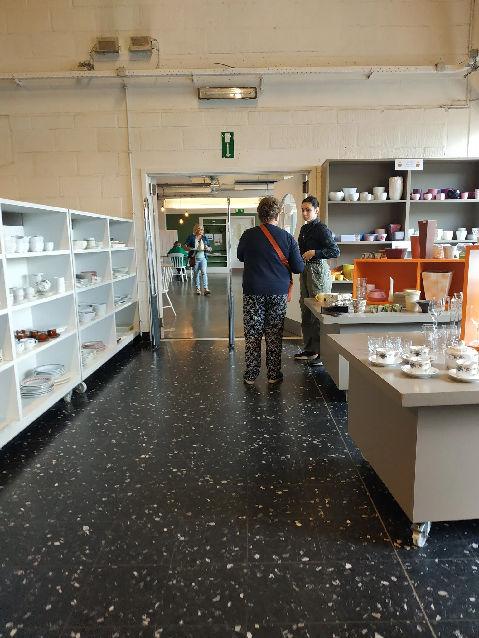 Winkelinterieur met witte stellingen vol serviesgoed, bruine kastjes en twee klanten die rondkijken