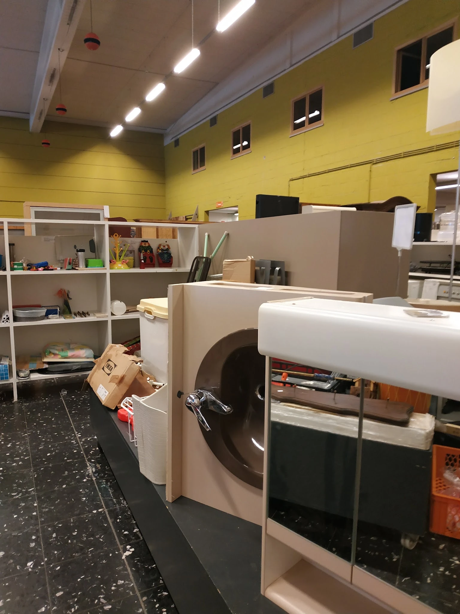 Winkelbinnenruimte met gele muren, stellingen met diverse spullen, grote witte apparaten en een zwarte vloer