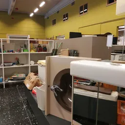 Winkelbinnenruimte met gele muren, stellingen met diverse spullen, grote witte apparaten en een zwarte vloer