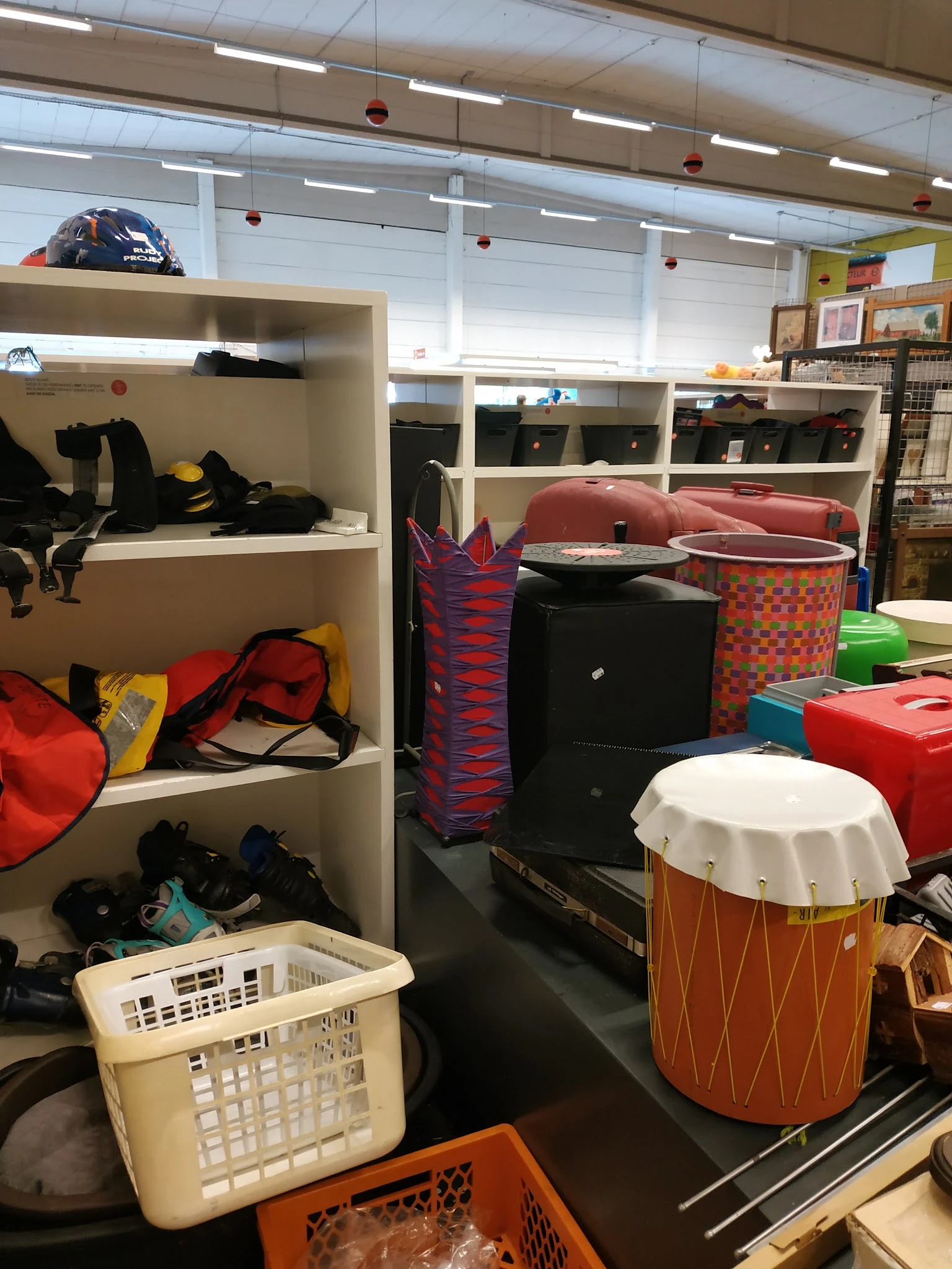 Stellingen met tweedehands spullen: zwarte tassen, rode tas, muziekinstrumenten en diverse opbergboxen in verschillende kleur
