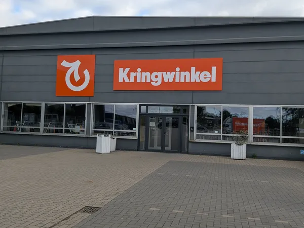 Grijze gevel van kringloopwinkel met oranje logo en winkelnaam, grote ramen en ingang met parkeerplaats ervoor