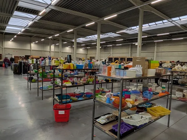 Overzicht van de winkelhal met tafels vol tweedehands spullen, dozen en artikelen onder industriële verlichting