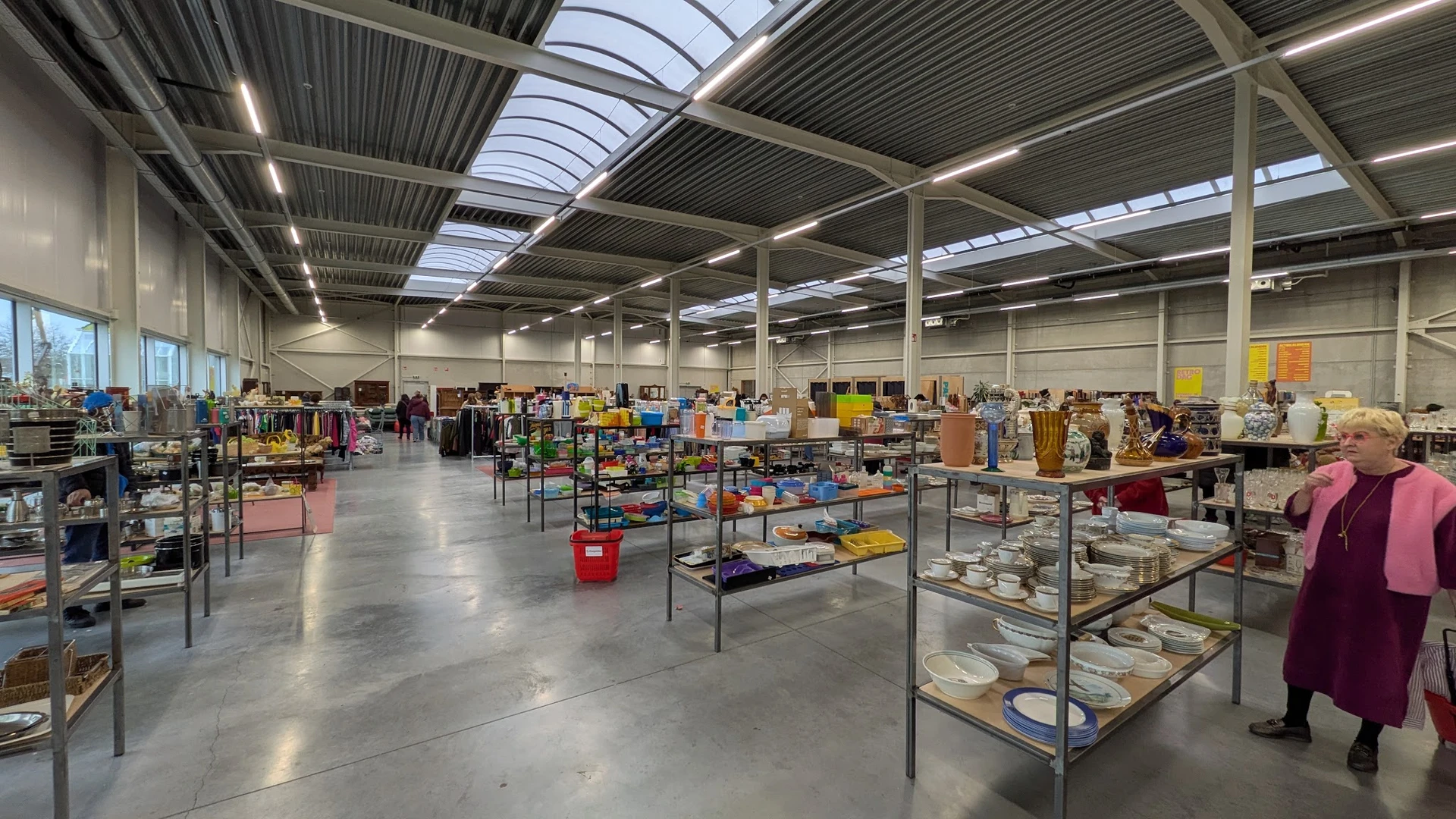Ruim magazijn met metalen stellingen vol tweedehands artikelen, onder daglichtdaken