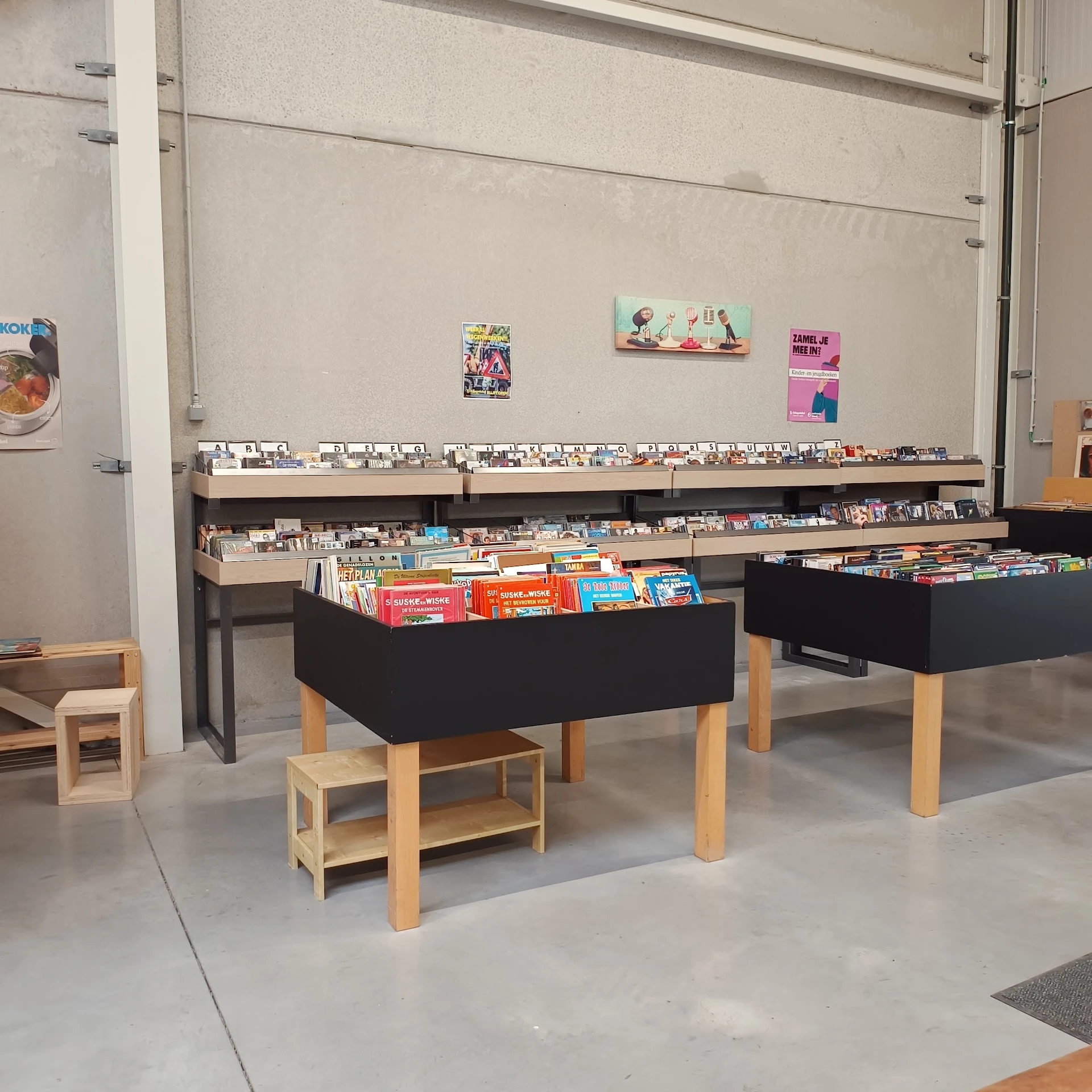 Zwarte display-tafels met houten poten gevuld met boeken, dvd's en media. Op de achtergrond stellingen met meer tweedehands a