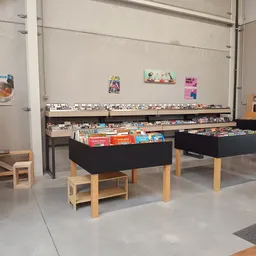 Zwarte display-tafels met houten poten gevuld met boeken, dvd's en media. Op de achtergrond stellingen met meer tweedehands a