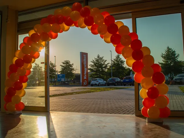 Ingang van kringloopwinkel met rode en gele ballonnen decoratie boven deuropening, uitzicht op parkeerplaats