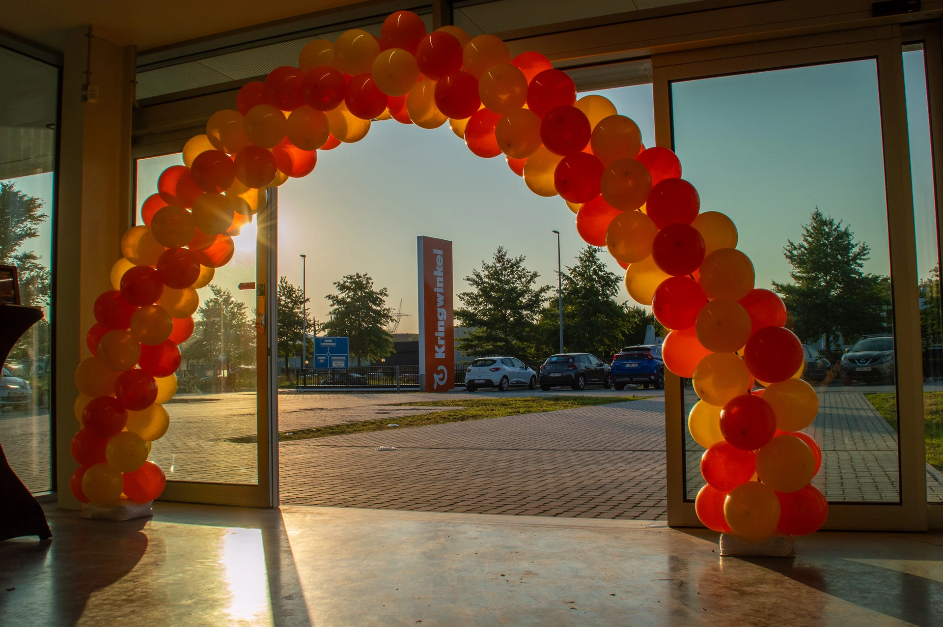 Ingang van kringloopwinkel met rode en gele ballonnen decoratie boven deuropening, uitzicht op parkeerplaats
