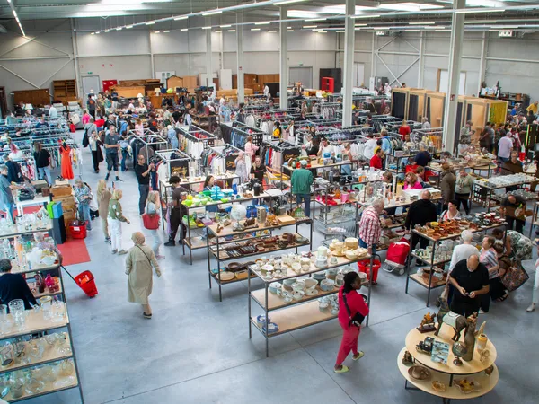 Overzicht van de kringloopwinkel met tafels vol diverse tweedehands artikelen, klanten die rondkijken en winkelende bezoekers