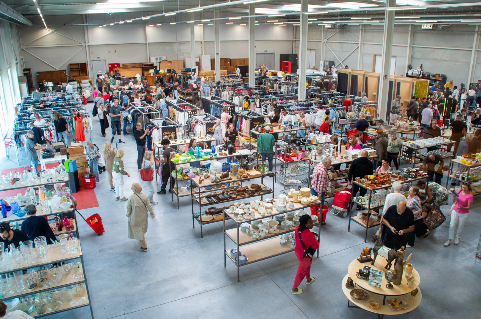 Overzicht van de kringloopwinkel met tafels vol diverse tweedehands artikelen, klanten die rondkijken en winkelende bezoekers