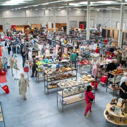 Overzicht van de kringloopwinkel met tafels vol diverse tweedehands artikelen, klanten die rondkijken en winkelende bezoekers