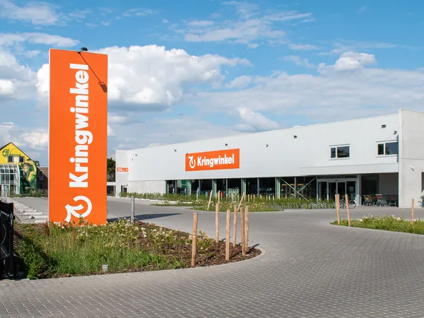 Moderne witte kringloopwinkel met groot oranje bord aan de voorkant, parkeerplaats en groene beplanting