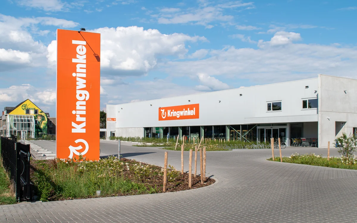 Moderne witte kringloopwinkel met groot oranje bord aan de voorkant, parkeerplaats en groene beplanting