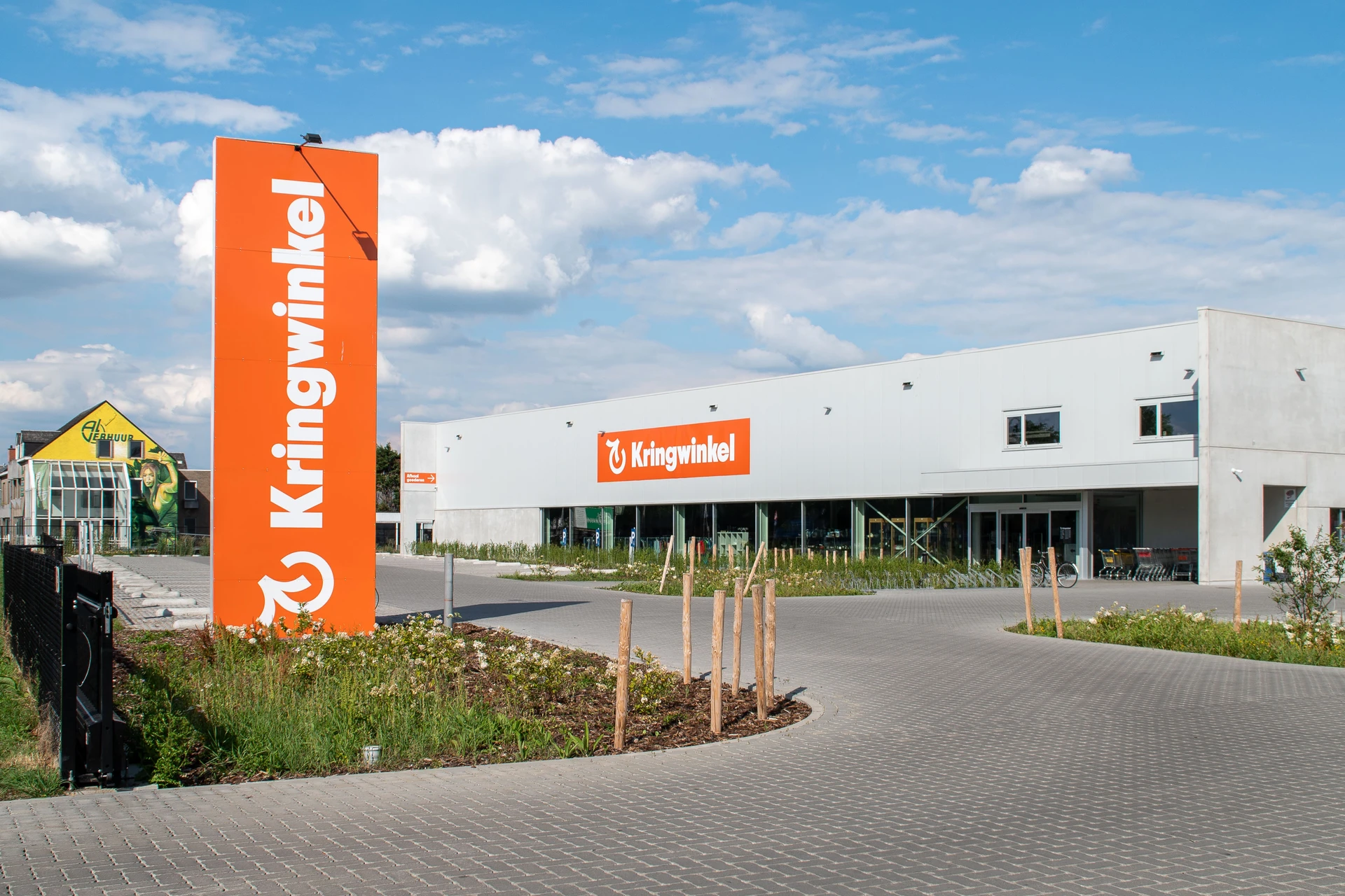 Moderne witte kringloopwinkel met groot oranje bord aan de voorkant, parkeerplaats en groene beplanting