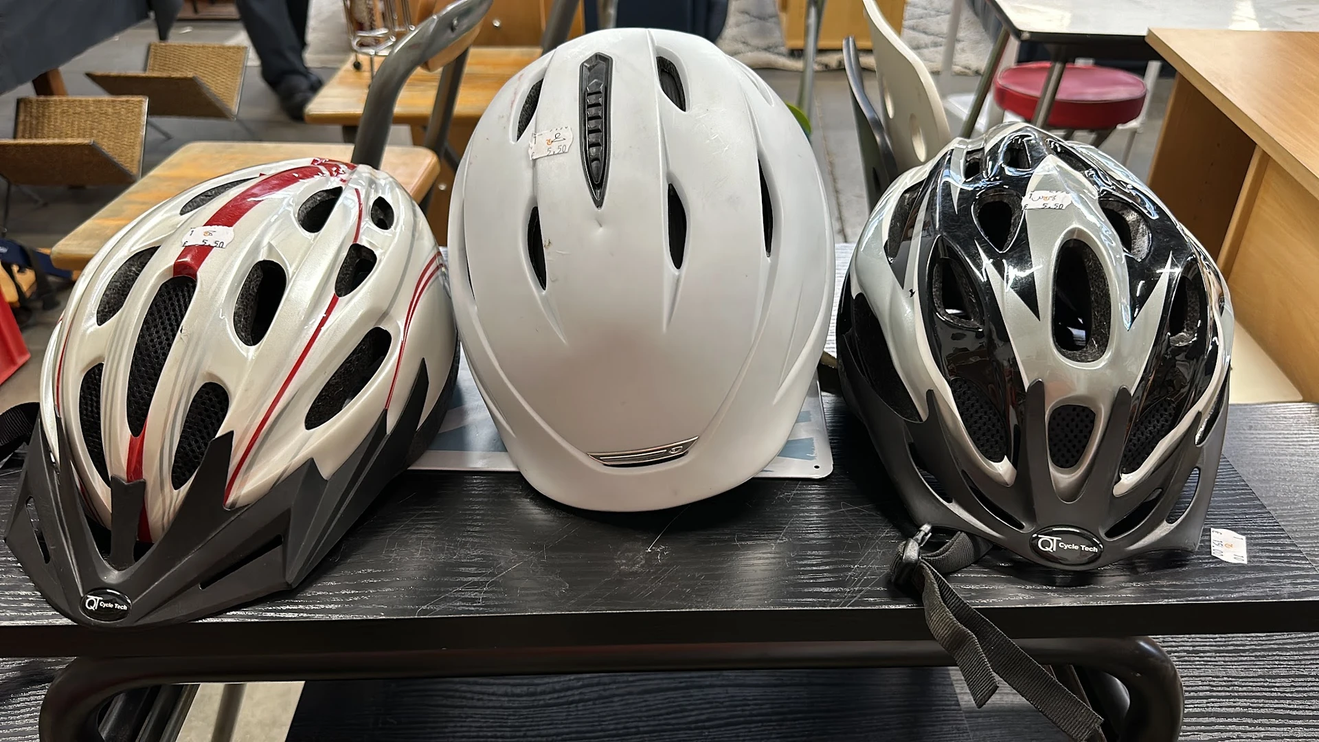 Drie fietshelmen in verschillende kleuren (wit met rood, wit, zwart-wit) uitgestald op een zwarte plank