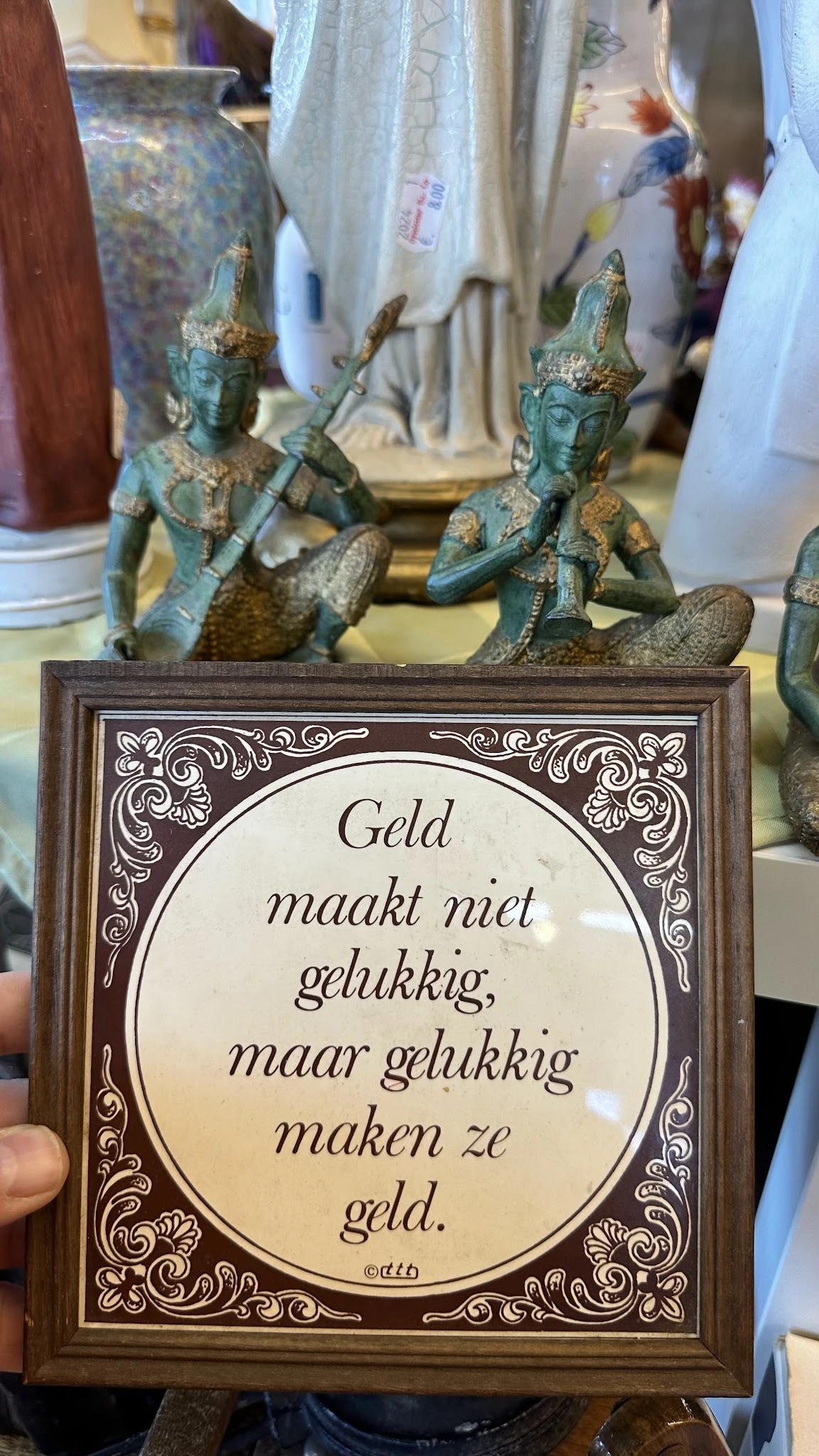 Close-up van een decoratief tegeltje met Nederlandse tekst 'Geld maakt niet gelukkig' in een bruine houten lijst, met decorat