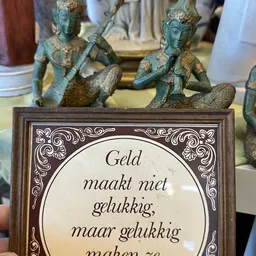 Close-up van een decoratief tegeltje met Nederlandse tekst 'Geld maakt niet gelukkig' in een bruine houten lijst, met decorat