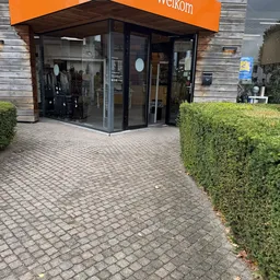 Winkelentree met glazen deuren en ramen, omgeven door groene hagen en bakstenen bestrating