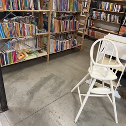 Binnenaanzicht van kringloopwinkel met houten boekenkasten vol DVD's en boeken, witte kinderstoel op voorgrond, betonnen vloe